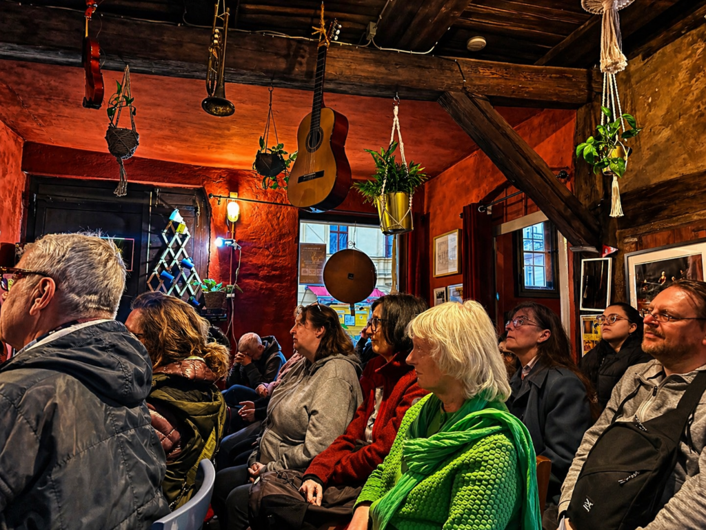 Besucher sitzen während des 2. Atelierabends im Atelier der Sinne zwischen roten Lehmwänden, Instrumenten und historischen Holzbalken.